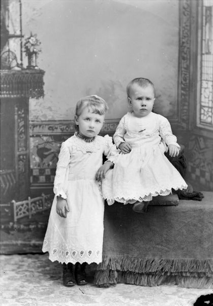 Studio portrait in front of a painted backdrop of two unidentified European American children. On the left, a young girl is standing on the with her left arm resting on a draped platform. She is wearing a short light-colored dress with lace trim, collar pin, and necklace. On the right a young girl is sitting on a small chair on the draped platform. She is wearing a light-colored dress with lace trim and a collar pin.