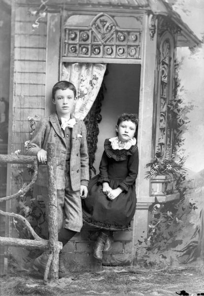 Studio portrait in front of a painted backdrop of two European American children. The boy is posing standing on the left with his left leg crossed over his right, and he has his right hand on a prop wooden fence. He is wearing light-colored knickerbockers, vest, suitcoat, and a ribbon bow tie. The girl is posing sitting in the center in a prop windowsill. She is wearing a dark-colored dress with a light-colored collar.