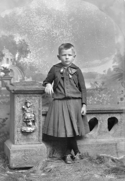 Studio portrait in front of a painted backdrop of an unidentified child posing standing in front of a prop stone wall with his right arm resting on the wall and his left leg crossed over their right leg. The child is wearing a dark-colored skirt, laced sweater, and ribbon bow tie.