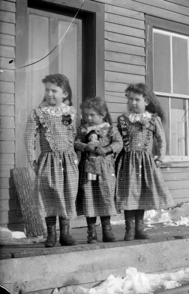Outdoor group portrait of three unidentified European American girls posing standing outdoors on a wooden sidewalk in front of a wooden building and door. All of the girls are wearing dresses made from the same light-colored plaid pattern fabric, and the girl in the middle is holding a doll.