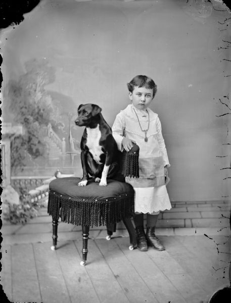 Studio portrait in front of a painted backdrop of an unidentified European American girl with short hair posing standing with her left arm on the back of a tasseled chair. Sitting on the chair is a dark-colored dog with a white chest. The girl is standing and holding onto the back of the chair and is wearing a light-colored dress and necklace.