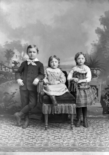 Studio portrait in front of a painted backdrop of an unidentified European American boy, standing on the left, and two girls. The youngest girl is sitting in a chair in the center, a the other girl is standing next to the chair on the right.