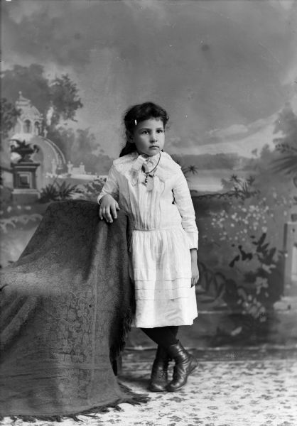 Studio portrait in front of a painted backdrop of an unidentified European American girl. She is wearing a light-colored dress and chain necklace, and is posing standing next to a draped chair.