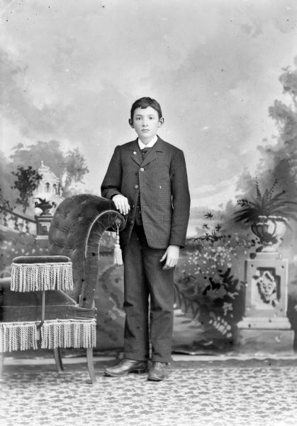 Full-length studio portrait in front of a painted backdrop of an unidentified European American boy standing with his arm resting on the back of an upholstered chair. He is wearing a suit with a lapel pin.