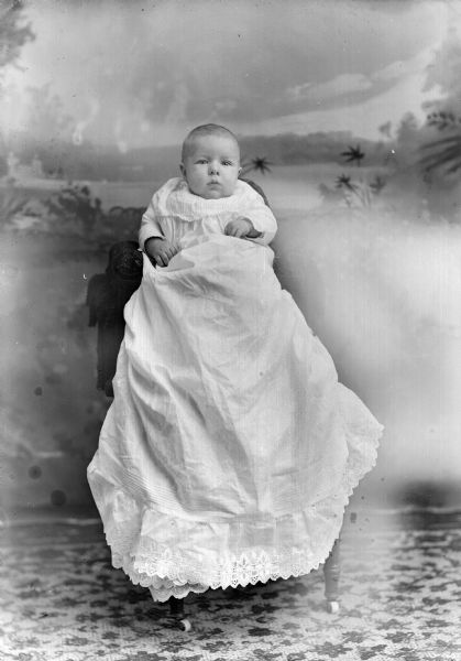 Studio portrait in front of an painted backdrop of an unidentified European American infant wearing a long christening gown.