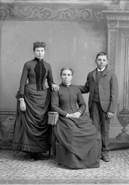 Studio portrait of an unidentified European American woman posing sitting in the center. She is flanked by a woman posing standing on the left, and an unidentified  boy posing standing on the right. The women are wearing dark-colored dresses with button down bodices and collar pins, and the boy is wearing a suit and necktie.