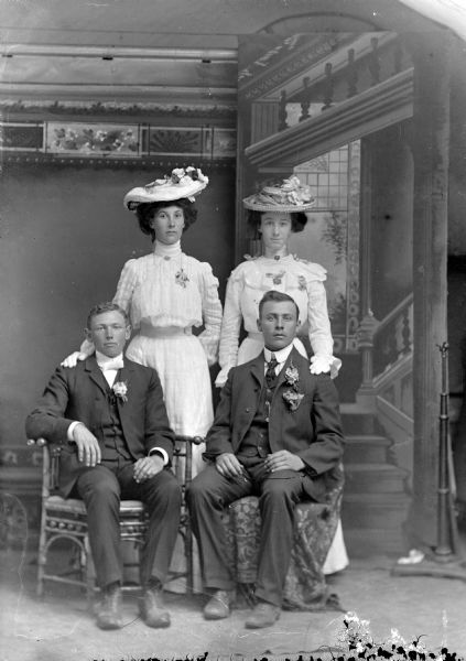 Studio portrait in front of a painted backdrop of two unidentified European American men posing sitting in front of two unidentified European American women posing standing. The men are wearing dark suits and corsages. The women are wearing light-colored dresses and flowered hats.
