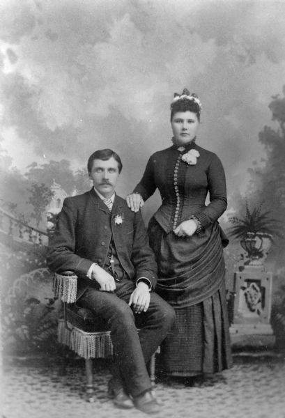 Studio portrait of an unidentified European American man posing sitting on the left, and an unidentified European American woman posing standing on the right, with her hand on the shoulder of the man. He is wearing a dark suit with a boutonnière. She is wearing a dark-colored dress with a button-down bodice and a corsage.
