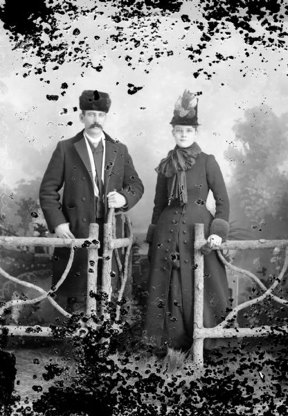 Studio portrait in front of a painted backdrop of Mr. and Mrs John Breirg, posing standing near a prop fence and gate. The man is standing on the left wearing a heavy coat and fur hat, and the woman is standing on the right and is wearing a heavy coat and flowered hat.