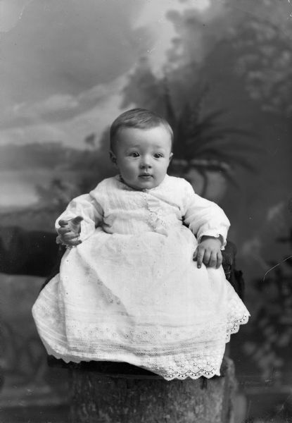 Studio portrait in front of a painted backdrop of an unidentified infant wearing a white christening gown posing sitting on a chair, which is on top of a prop tree trunk.
