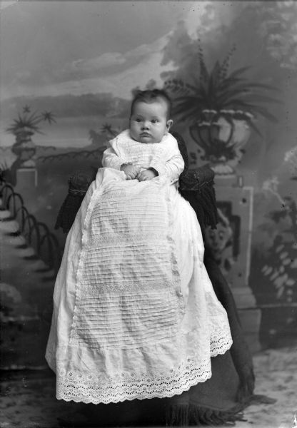 Studio portrait in front of a painted backdrop of an unidentified infant posing sitting in a chair, which is on a cloth-covered stand. The infant is wearing a long, white christening gown.