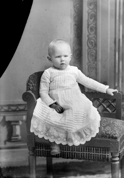 Studio portrait in front of a painted backdrop of an unidentified child wearing a white gown posing sitting on a settee.