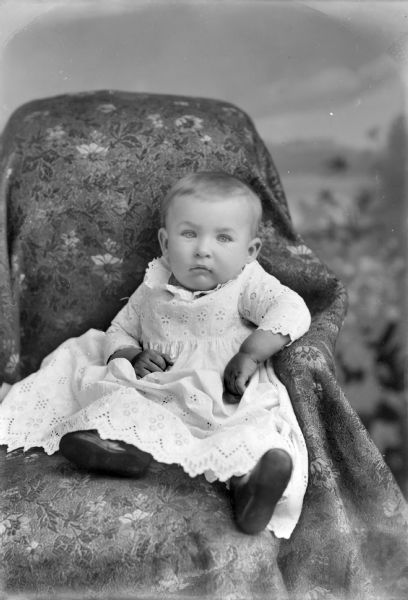 Studio portrait in front of a painted backdrop of an unidentified infant in a white gown with eyelets, posing sitting in a chair draped with a cloth.