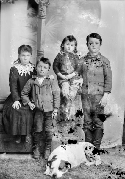 Studio portrait in front of a painted backdrop of four unidentified children. A girl is posing sitting on the left and has her arm around a boy posing standing next to her. The smallest child is sitting higher up on a prop stone column, and next to him on the right is an older boy posing standing. A dog is lying on the floor in the foreground.