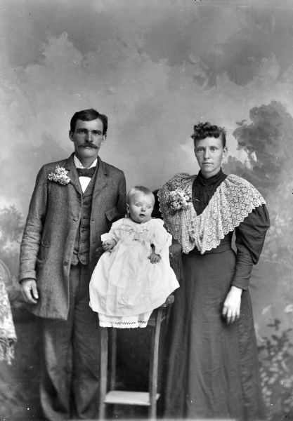 Three-quarter length studio portrait in front of a painted backdrop of an unidentified family. An infant is posing sitting in the center on a high chair, wearing a long white gown. The infant is flanked by a man with a moustache posing standing on the left, and a woman with a lace collar and a corsage posing standing on the right.