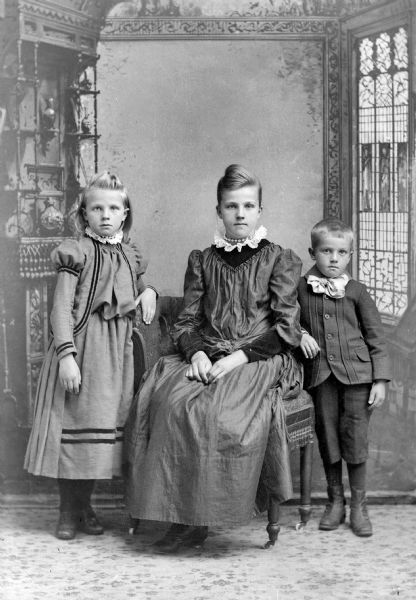 Studio portrait in front of a painted backdrop of an unidentified family. A young woman is posing sitting in the center, and is flanked by a girl posing standing on the left, and a boy posing standing on the right.
