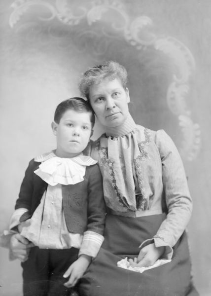 Three-quarter length studio portrait in front of a painted backdrop of a boy posing standing on the left, and a woman posing sitting on the right. The woman has her arm around the boy.