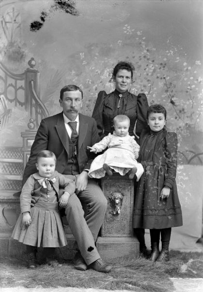 Studio group portrait in front of a painted backdrop of an unidentified family. A man and an infant are posing sitting on a prop stone wall and column. On the left a boy is standing and leaning on the knee of the man. A girl on the right is standing next to the infant, and a woman is standing behind her.
