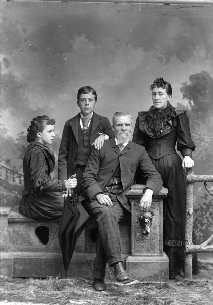 Studio portrait in front of a painted backdrop of an unidentified family. A man is posing sitting on a prop stone wall with his elbow resting on a stone column. A young woman with an umbrella is sitting sideways on the stone wall next to him. Behind her a boy is posing standing with his hand on the man's shoulder. On the right is a woman posing standing with her hand on a prop wooden fence.