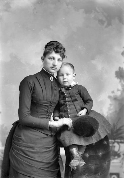 Studio portrait in front of a painted backdrop of an unidentified woman and girl. The woman is wearing a cameo at the throat of her dress which has a laced bodice. She is posing standing on the left, holding a girl posing sitting on the back of an overstuffed chair on the right. The girl is holding a hat.