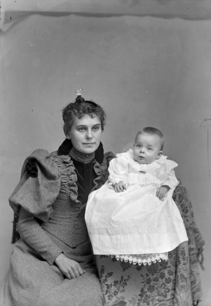 Studio portrait of an unidentified woman and infant. The woman is wearing a jeweled comb in her hair and is posing sitting and holding an infant who is posing sitting in a raised, cloth-draped chair on the right.