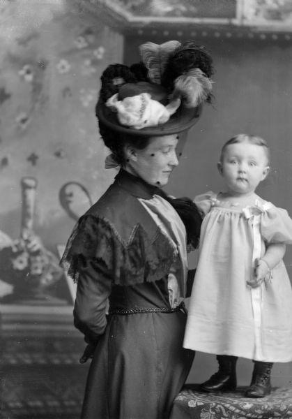 Studio portrait in front of a painted backdrop of unidentified family. A woman wearing a feathered hat is posing in profile standing on the left. She is holding a girl posing standing on a cloth-covered table on the right.