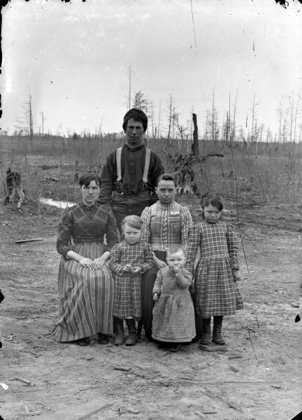 Outdoor group portrait of unidentified family. Two women are posing sitting in front of a man posing standing, with three girls posing standing. Location identified as the east side of town.