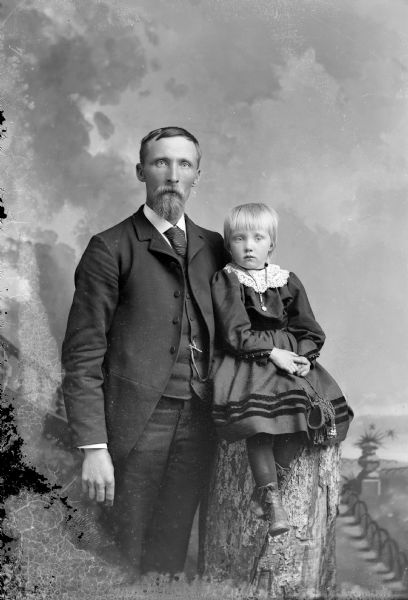 Studio portrait in front of a painted backdrop of an unidentified family. A man is posing standing on the left, with his arm around a girl posing sitting on a prop stump on the right. The girl is wearing a necklace and is holding a small beaded handbag.
