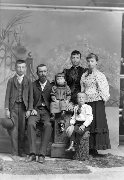 Studio portrait in front of a painted backdrop of an unidentified family. A man and woman are posing with a young girl and boy, and an older girl and boy. The group is sitting and standing near a faux stone balustrade, and the young boy is sitting on a tree stump. The edge of the backdrop is on the right.