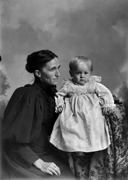 Studio portrait in front of a painted backdrop of an unidentified family. A woman is sitting on the left and holding a child, who is posing sitting on a draped chair on the right. The woman has a ring on her finger and is wearing a dark dress with dark lace bow at her throat. The child is wearing a long white dress with white lace collar.