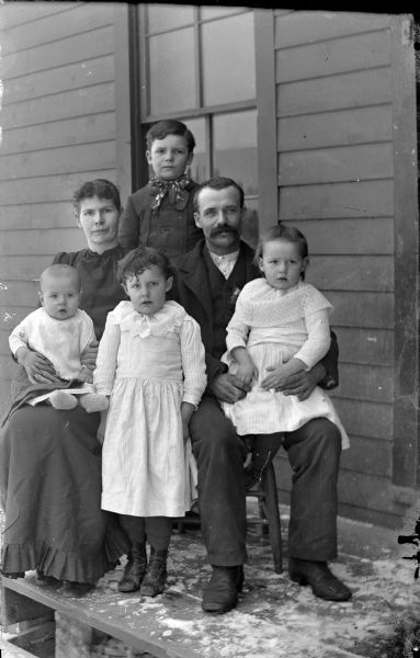 Outdoor group portrait of an unidentified family. A man and woman are posing sitting, both holding a child, with a girl posing standing between them, and a boy is standing behind them. There is snow on the stoop.