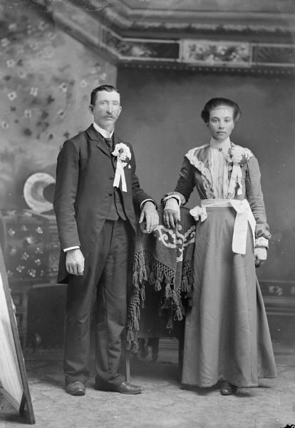 Studio portrait in front of a painted backdrop of an unidentified man posing standing on the left and a woman is posing standing on the right.
