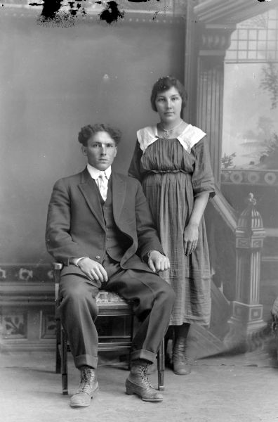 Studio portrait in front of a painted backdrop of an unidentified man posing sitting on the left and a woman posing standing on the right.