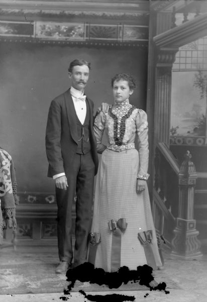 Full-length studio portrait in front of a painted backdrop of an unidentified man posing standing on the left and an unidentified woman posing standing on the right.