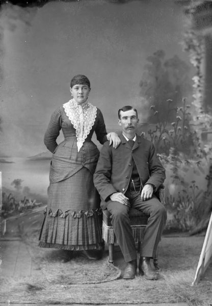 Full-length studio portrait in front of a painted backdrop of an unidentified woman posing standing on the left and an unidentified man posing sitting on the right.