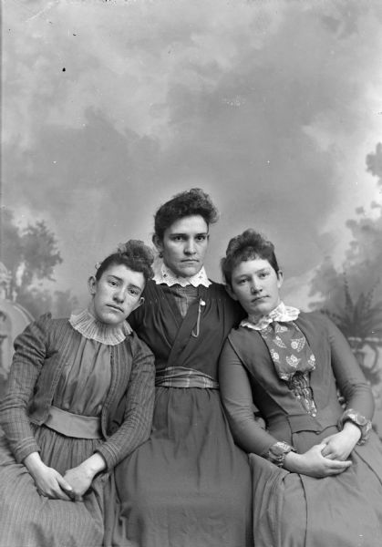 Three-quarter-length studio portrait in front of a painted backdrop of two unidentified young women posing sitting on the left and right, flanking an older unidentified woman posing sitting in the center. Perhaps a mother and her two teenage daughters.