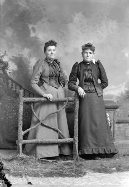 Full-length studio portrait in front of a painted backdrop of an unidentified woman posing standing behind a wooden fence on the left, and an unidentified woman posing standing on the right.
