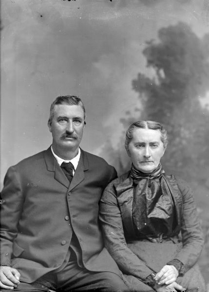 Waist-up studio portrait in front of a painted backdrop of an unidentified middle-aged man posing sitting on the left, and an unidentified middle-aged woman posing sitting on the right.