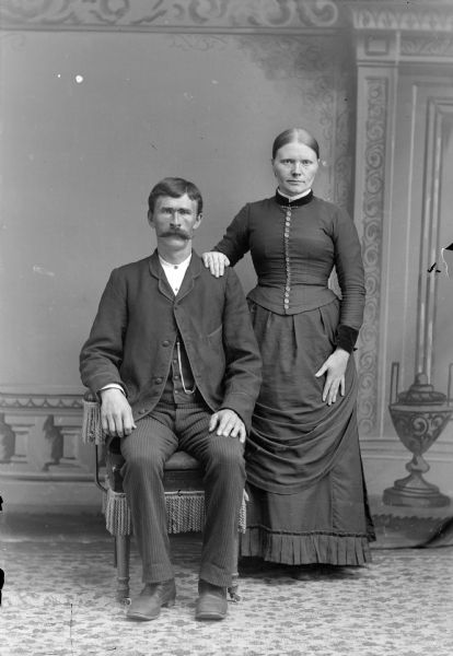 Full-length studio portrait in front of a painted backdrop of an unidentified man with a moustache posing sitting on the left, and an unidentified woman posing standing on the right.