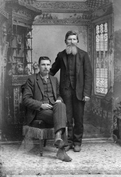 Full-length studio portrait in front of a painted backdrop of an unidentified man with a goatee posing sitting on the left, and an unidentified man with a full beard posing standing on the right.