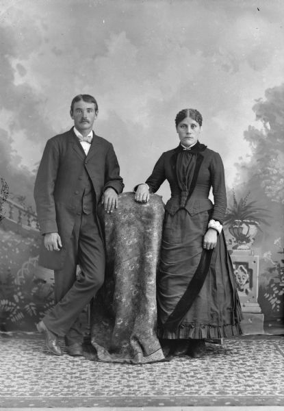 Full-length studio portrait in front of a painted backdrop of an unidentified man posing standing on the left, and an unidentified woman posing standing on the right. Both of them are wearing wedding rings and are resting their arms on a cloth-covered table.