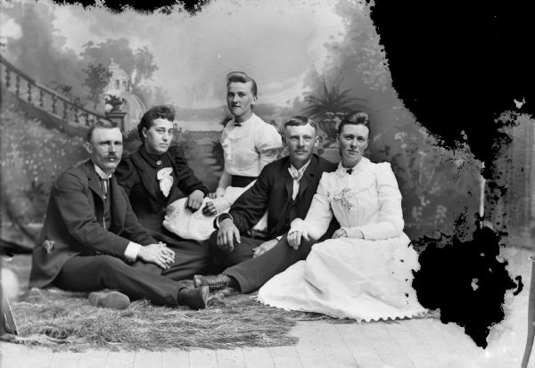 Studio group portrait of men and women sitting on the floor in front of a painted backdrop. From left is a man, then two women in the left center and center, and a man and woman on the right.