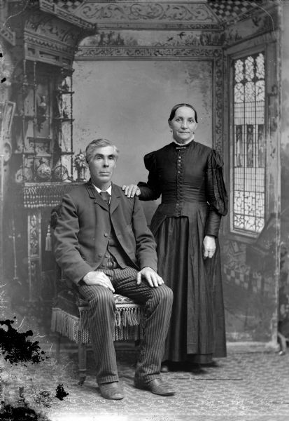Full-length studio portrait in front of a painted backdrop of an unidentified man posing sitting on the left, and an unidentified European American woman, with her hand on the man's shoulder, posing standing on the right.
