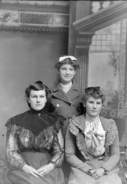 Studio Portrait of two unidentified women posing sitting in front of a woman posing standing in the center behind them.
