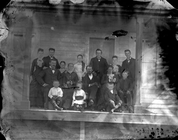 Outdoor group portrait of an unidentified group of men, women, and children, posing sitting and standing on the porch of a wooden building. It's noted on the envelope that the woman seated left of center has a goiter.