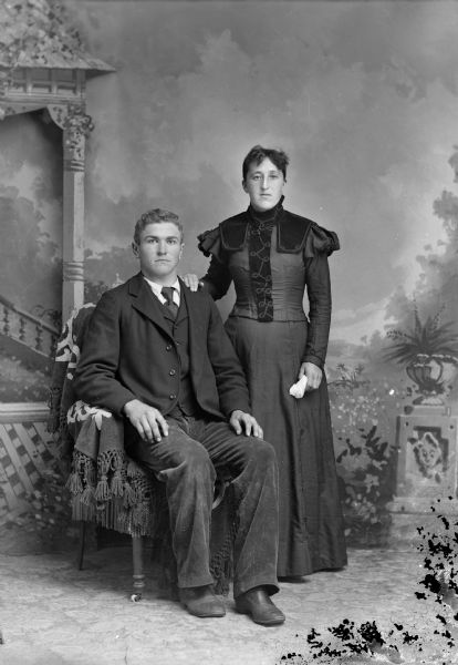 Studio portrait in front of a painted backdrop of a man posing sitting on the left, and a woman posing standing on the right.