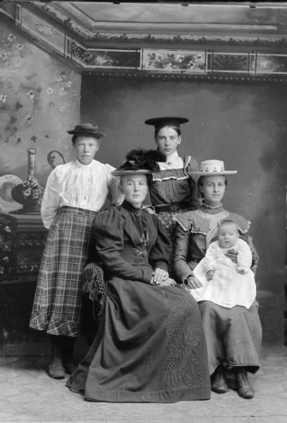 Studio group portrait in front of a painted backdrop of two women posing sitting. The woman on the right is holding an infant. Behind them are two girls posing standing. All of the women are wearing hats.
