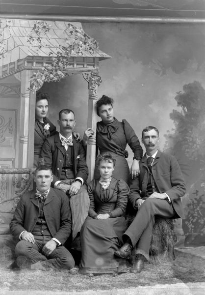 Studio group portrait with prop porch setting of three men and a woman posing sitting, with two women posing standing in front of a painted backdrop.