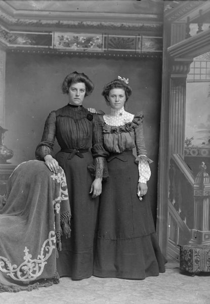 Studio portrait n front of a painted backdrop of a woman posing standing on the left and a woman posing standing on the right.