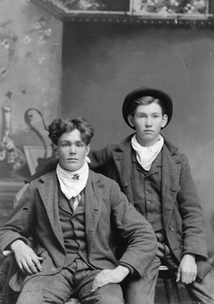 Three-quarter length portrait of two young men wearing neckerchiefs posing sitting together in front of a painted backdrop.